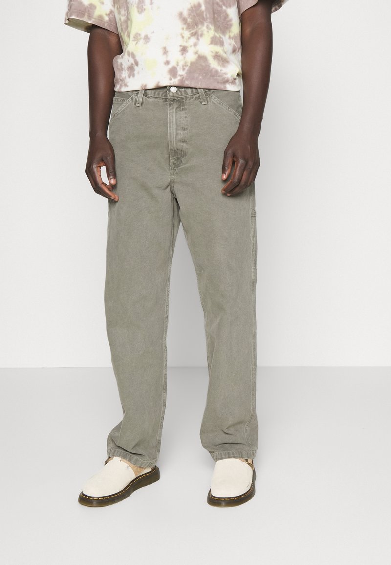 Personne portant un pantalon droit vert clair, des chaussures blanches sans lacets et un t-shirt à manches courtes beige et marron tie-dye, debout devant un fond blanc.