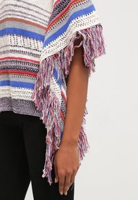 Poncho lavorato a maglia con strisce multicolori, caratterizzato da bordi frangiati in diverse tonalità. Leggero, con un design aperto e una finitura testurizzata.