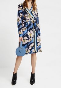 Robe portefeuille florale à manches longues en bleu marine et couleurs pastel, avec une taille à nouer et un sac à main rond bleu. Bottines noires incluses.