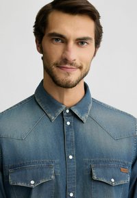 Camisa de denim azul con cuello puntiagudo, dos bolsillos en el pecho y botones a presión. Notable desgastado claro y detalle de costura en contraste.
