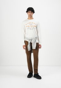 Jonge man die staat in een wit overhemd met lange mouwen met oranje tekst, bruine broek, grijze trui om zijn middel geknoopt, en zwarte schoenen tegen een effen achtergrond.