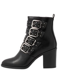 Botín de cuero negro hasta el tobillo con punta afilada, que presenta cuatro grandes hebillas de plata y acentos de tachuelas en las correas. Tacón ancho.