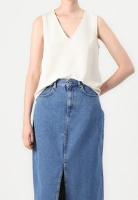 Haut sans manches beige clair avec un décolleté en V, associé à une jupe en denim bleu taille haute, longueur cheville, avec une fente frontale et des coutures classiques.