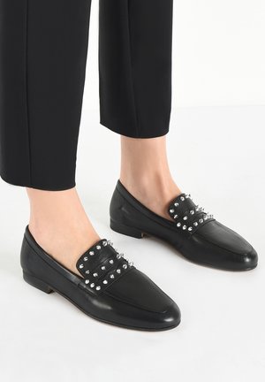 Mocassins en cuir noir à bouts pointus, ornés de pointes argentées sur la partie supérieure. Talon bas et texture lisse. Portés avec un pantalon noir raccourci.