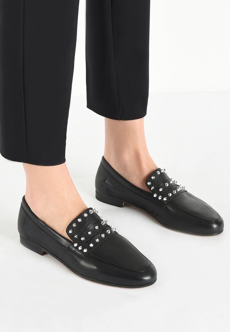 Mocassins en cuir noir à bouts pointus, ornés de pointes argentées sur la partie supérieure. Talon bas et texture lisse. Portés avec un pantalon noir raccourci.
