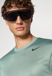Czarne okulary przeciwsłoneczne o zakrzywionym kształcie, noszone z miętową koszulą z krótkim rękawem, na której piersi znajduje się małe czarne logo Nike w kształcie pukla.