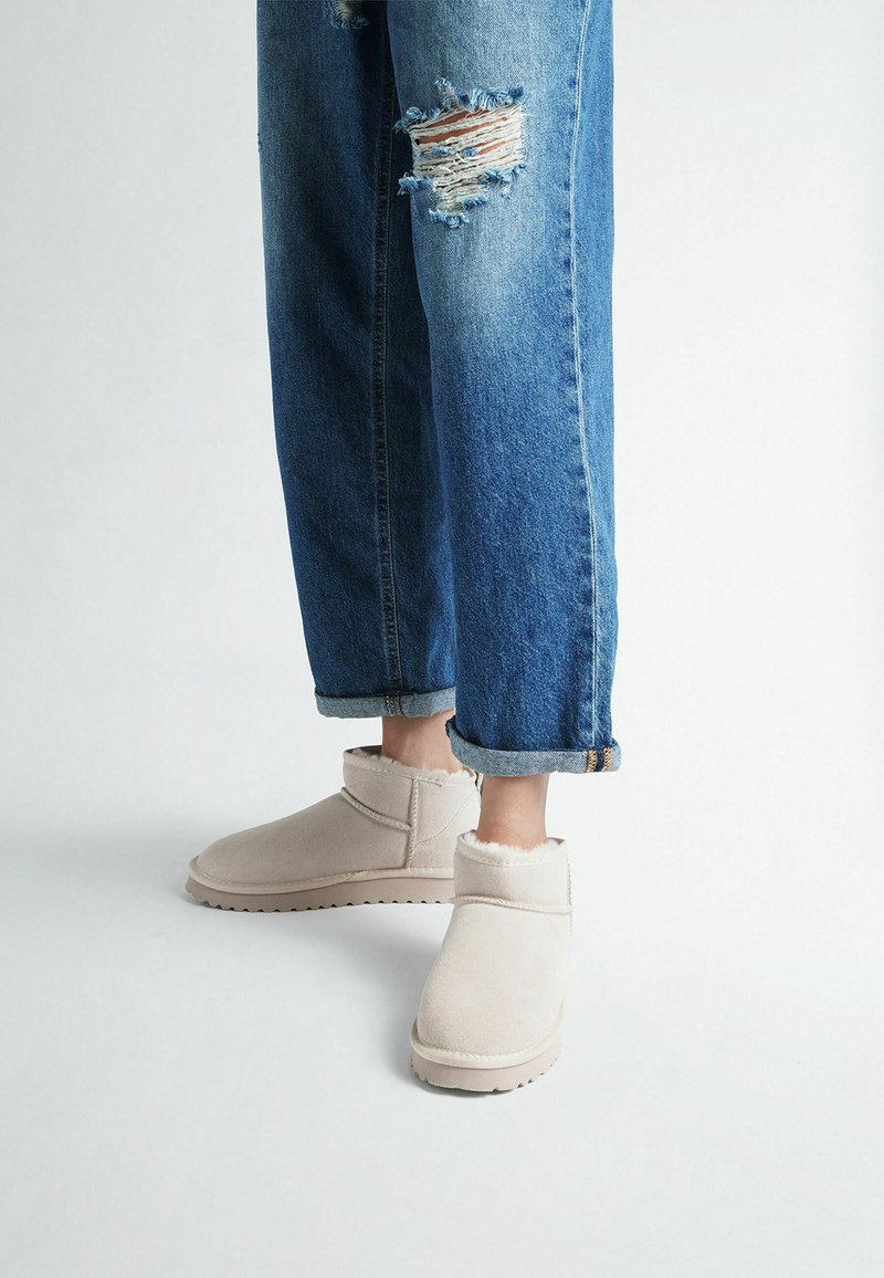 Persona che indossa jeans blu strappati e arrotolati e stivaletti beige foderati in pile su uno sfondo chiaro.
