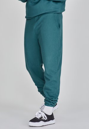 Pantalones de chándal verde azulado con puños elásticos, bolsillos laterales y una textura suave, combinados con zapatillas negras con cordones y calcetines blancos de caña corta.