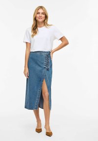 Maxirock aus Denim mit seitlichem Schlitz und Schnürdetail in der Taille. Kombiniert mit einem schlichten weißen T-Shirt und braunen Schuhen mit Blockabsatz.