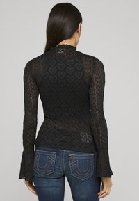 Top in pizzo nero con motivi a cuore, maniche lunghe con polsini svasati e collo alto. Indossato sopra jeans blu scuro con cuciture decorative.