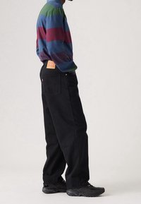 Svart denimjeans med en löst sittande passform, med en bakpatch med etikett och subtila sömdetaljer. Matchas med en flerfärgad randig långärmad skjorta.