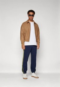 Bruine bomberjack, wit T-shirt, marineblauwe joggingbroek met groene zijstrepen en witte sneakers. Model draagt zwarte zonnebril.