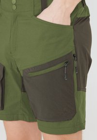 Cargo shorts i mörk och ljusgrön tyg, med flera fickor med dragkedjor och en strukturerad yta. Anmärkningsvärda sömdetaljer finns.