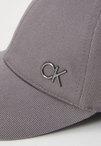 Grijze baseballpet van een textured stof, met een zwart metalen "CK" logo aan de voorkant. Gebogen klep, gestikte accenten.