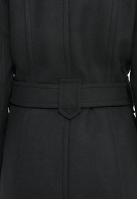 Cappotto nero con cintura, realizzato in tessuto testurizzato, presenta cuciture verticali e una fibbia a punta. Il design sottolinea una silhouette strutturata.