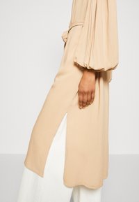 Beżowa sukienka z długim rękawem, posiadająca bufiaste rękawy i boczny rozcięcie. Wykonana z gładkiego materiału, z wiązanym paskiem w talii.