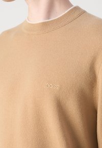 Zoom su una persona che indossa un maglione lavorato a maglia beige con collo a girocollo a coste e un piccolo logo ricamato "BOSS" sul petto.