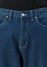 Zili džinsi no zila denima ar sudraba pogu aizdari, jostas cilpām un redzamām priekšējām kabatām uz melna fona.