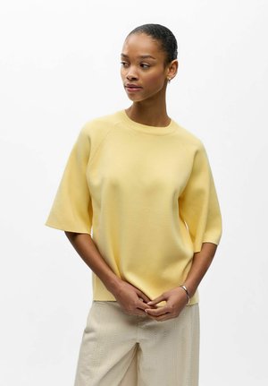 Femme aux cheveux attachés, portant un pull jaune à manches courtes et un pantalon beige clair, regardant vers la gauche avec les mains légèrement jointes.