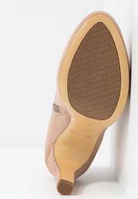 Chaussure à talon haut en daim beige, présentée en angle pour montrer la semelle en caoutchouc texturée et le petit embout de talon sur fond blanc.