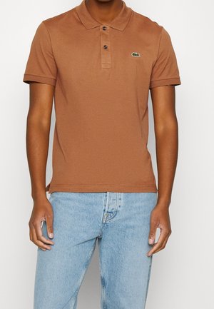 Mężczyzna w brązowej koszulce polo z krótkim rękawem z małym zielonym logo na piersi oraz w jasnoniebieskich dżinsach.