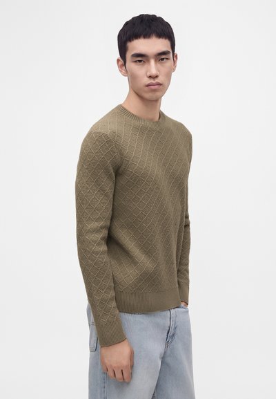 Jeune homme portant un pull texturé vert olive et un jean bleu clair, debout devant un fond blanc uni.