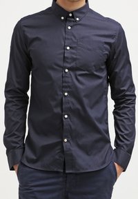 Námornícky modrá košile s dlouhým rukávem na knoflíky, vyrobená z hladké bavlny, s kapsou na hrudi a bílými knoflíky podél přední légy.