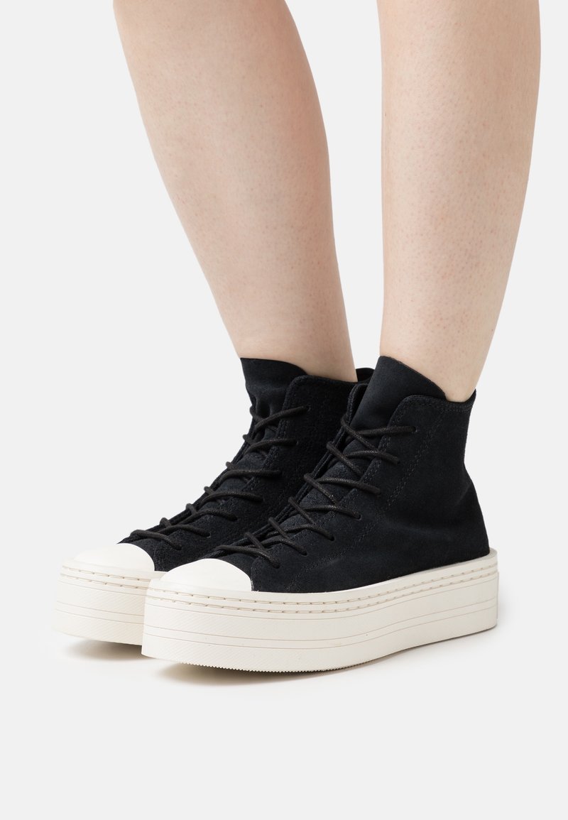 Paire de baskets montantes noires à plateforme avec semelles épaisses blanches portées sur des jambes nues sur fond blanc.