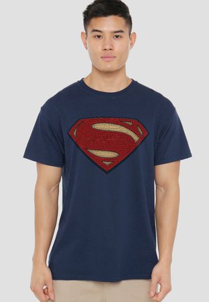 Young man standing facing forward wearing a navy blue t-shirt with a large textured red and gold Superman logo.