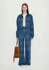 Casaco de ganga azul e jeans de perna larga coordenados com um padrão texturizado. A segurar uma mala de couro castanha em forma de balde. Top branco por baixo.