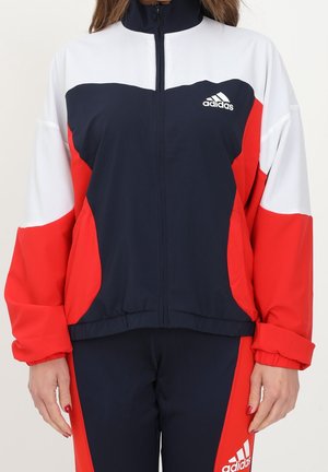 Vrouw draagt een marineblauw, rood en wit Adidas jack met rits en bijpassende broek met Adidas-logo op borst en dij.