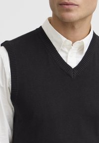 Svart stickad ärmlös tröja med V-ringning, med ribbade kanter och bärs över en vit skjorta med krage. Slät textur, minimalistisk design.