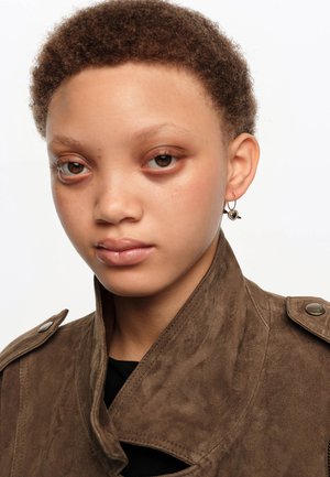 Joven con cabello corto y rizado que lleva una chaqueta de ante marrón y un pendiente en forma de corazón con una flecha en la oreja izquierda.