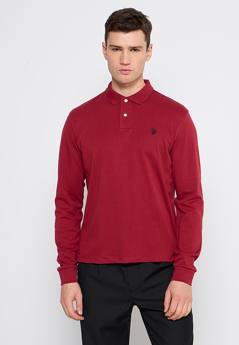 Homme portant un polo rouge à manches longues avec un petit logo noir sur la poitrine gauche et un pantalon noir, se tenant devant un fond blanc.