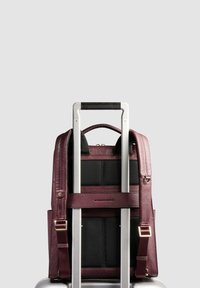 Zaino in pelle color marrone scuro con schienale imbottito in rete nera, spallacci regolabili e componenti metallici, montato sulla maniglia di una valigia argento.