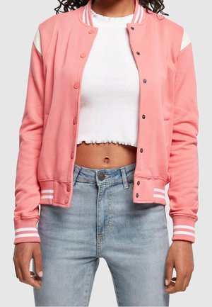 Vrouw die een roze varsityjack met drukknopen draagt over een wit cropped shirt en lichtblauwe high-waist jeans, staand met ontspannen armen.