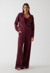 Vrouw in bordeauxrode wijde broek, bijpassende rits-hoodie en tanktop, staand met één hand in de zak tegen een effen achtergrond.