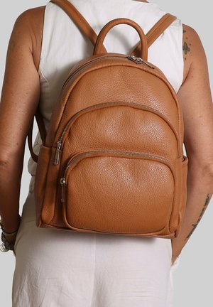 Donna che indossa un outfit bianco senza maniche con uno zaino in pelle color cuoio con molteplici scomparti con cerniera e una maniglia superiore.