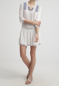 Vit klänning med mönstrad blå broderi, 3/4 ärmar, draperad midja och utsvängd kjol. Kombinerad med flata sandaler med remmar.