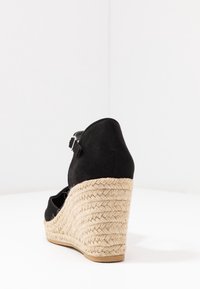 Schwarzer Wildlederwedge mit geflochtenem Jute-Absatz, geschlossener Ferse und verstellbarem Knöchelriemen. Flache Gummisohle für einen guten Halt.