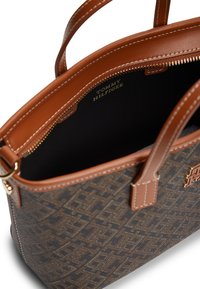 Καφέ και μαύρη τσάντα με σχέδιο, με δερμάτινες λαβές και περιμετρικά τμήματα σε ταμπά χρώμα, που διαθέτει χρυσό φερμουάρ και το λογότυπο "Tommy Hilfiger" τυπωμένο στο εσωτερικό.