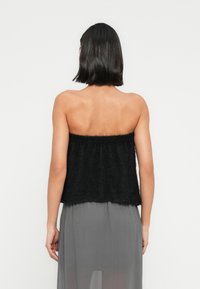 Γυναίκα με μαύρα μαλλιά μέχρι τους ώμους, που φοράει μαύρο τοπ χωρίς τιράντες και διαφανή γκρι φούστα, γυρισμένη προς τα πίσω σε απλό λευκό φόντο.