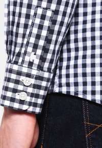 Cuff di una camicia a scacchi bianco e nero con due bottoni bianchi, indossata sopra un paio di jeans in denim blu scuro con cuciture arancioni visibili.