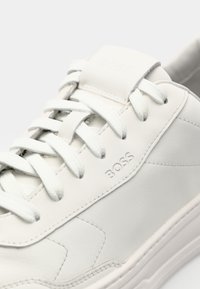 White leather sneaker with tonal stitching, flat laces, and a minimalistic design. Embossed logo on the side and smooth texture.