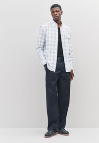 Camicia con bottoni a quadri azzurro chiaro e bianca con taschino sul petto, indossata sopra una maglietta nera, abbinata a pantaloni larghi scuri e scarpe nere.