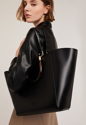Sac fourre-tout en cuir noir avec une forme structurée, présentant une finition brillante et des accents de poignées en plastique transparent. Détails en métal doré visibles.