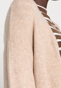 Jasnobeżowy sweter z miękkiego materiału o luźnym kroju, z wyraźnymi szwami na ramionach i subtelną fakturą. Pod spodem widoczne paski.