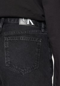 Vaqueros de mezclilla negros con un bolsillo trasero, una etiqueta de cuero en la cinturilla con "Calvin Klein Jeans" y un acabado suave y texturizado.