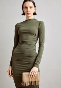 Długa sukienka w oliwkowej zieleni z marszczeniami. Modelka trzyma złotą kopertówkę z frędzlami. Gładka tkanina i dopasowany krój.