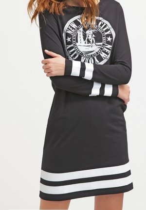 Donna che indossa un vestito nero a maniche lunghe con strisce bianche sui polsini e sull'orlo, con una grafica circolare "New York City" e uno skater.